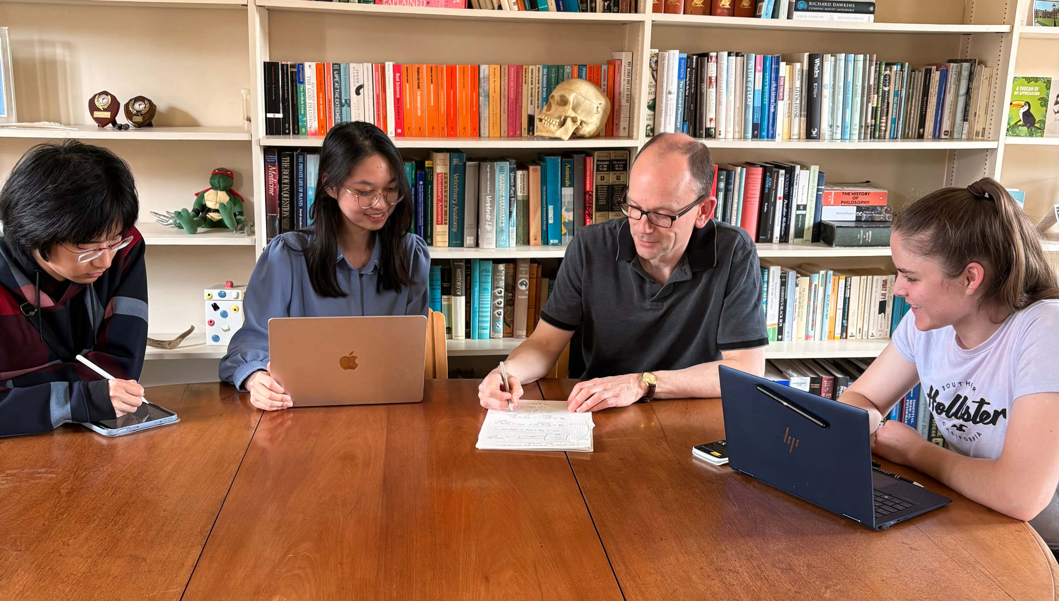 A group of students sitting at a large wooden table during a supervision session. They have laptops in front of them and the teacher is writing on a notepad while speaking to them. They are seated in front of shelves filled with books