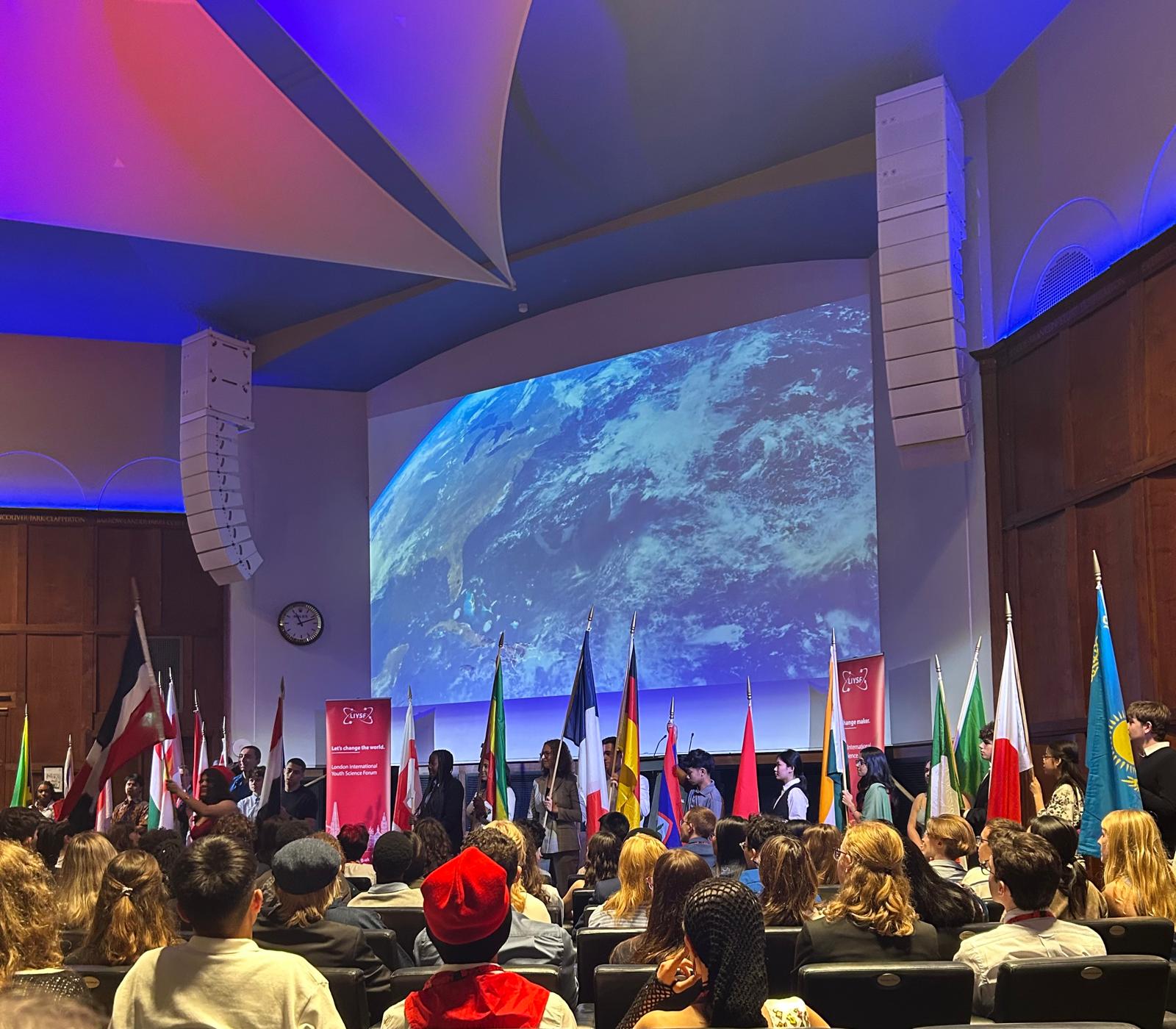 Audience at the LISYF opening ceremony, seated, with different national flags at the front of the auditorium. A large image of the earth is projected on to the wall behind the flag bearers. 