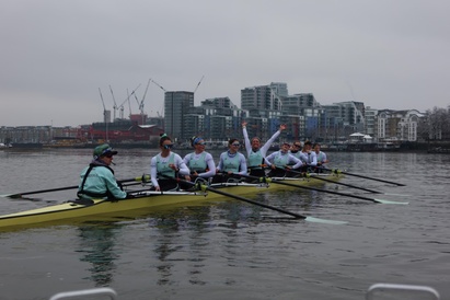A photograph of the Cambridge rowing crew seated in their boat on the river, smiling and laughing. Jenna Armstrong is seated and looking directly at the camera. She is smiling and raising both of her arms in celebration.