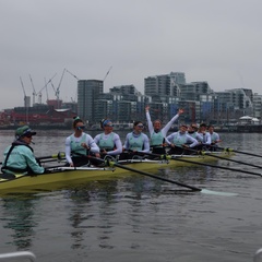 A photograph of the Cambridge rowing crew seated in their boat on the river, smiling and laughing. Jenna Armstrong is seated and looking directly at the camera. She is smiling and raising both of her arms in celebration.