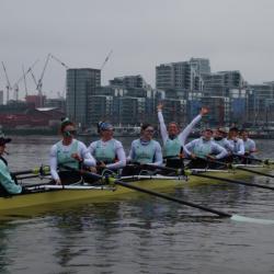 A photograph of the Cambridge rowing crew seated in their boat on the river, smiling and laughing. Jenna Armstrong is seated and looking directly at the camera. She is smiling and raising both of her arms in celebration.