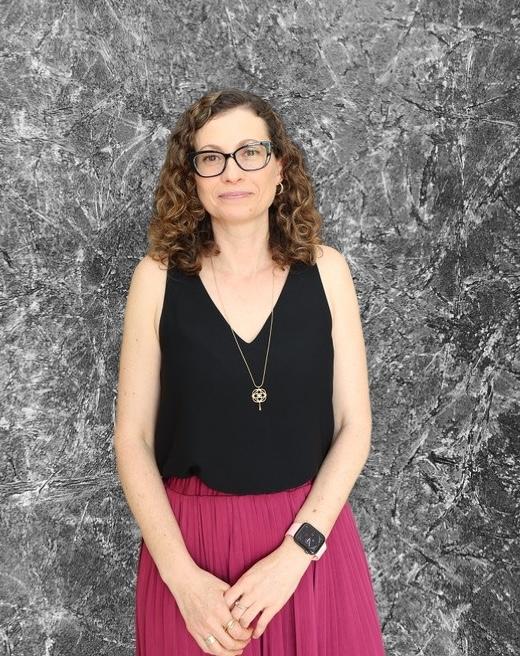 Dr Guela Hanin stands, facing the camera. She is standing in front of a grey background and she is smiling at the camera